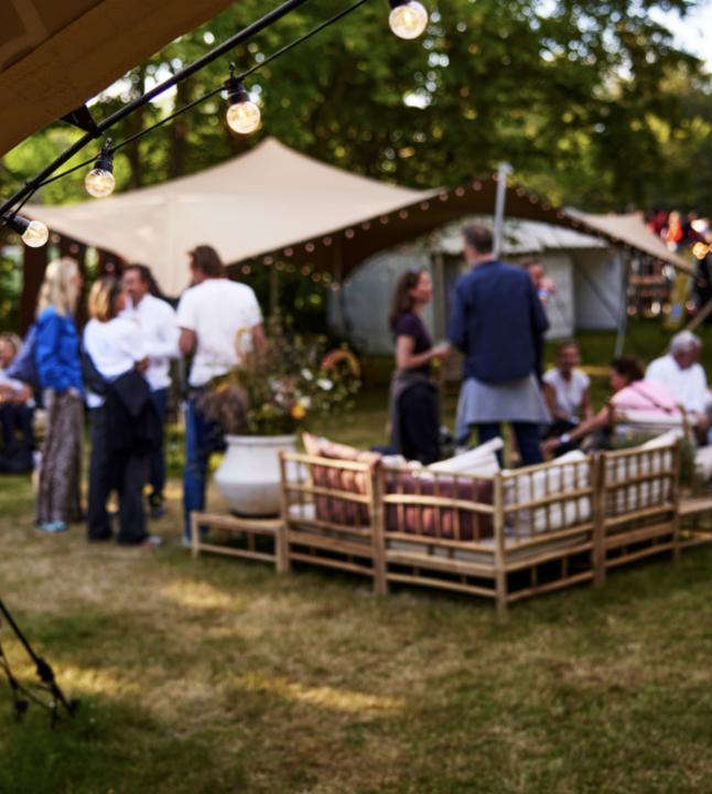 Gæster hygger sig i en afslappet lounge med bambusmøbler, lyskæder og sejltelte omgivet af grønne træer ved Heartland Festival.