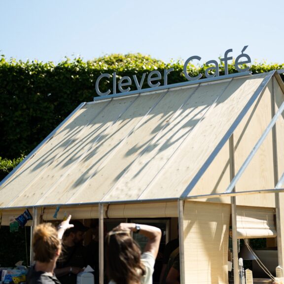 Blå himmel og høj grøn busk i baggrunden. Foran står Clever café med to mennesker der pejer mod caféen