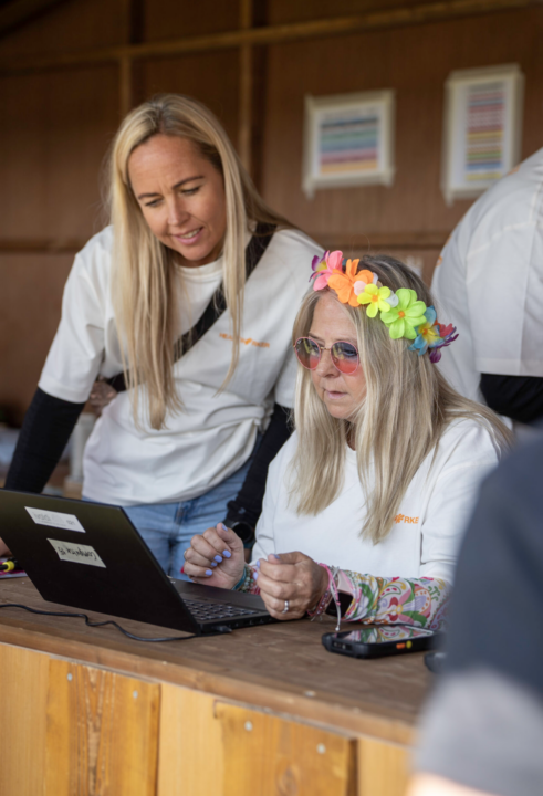 To frivillige i hvide t-shirts arbejder ved en bærbar computer, den ene med farverig blomsterkrans i håret og lyserøde solbriller, ved Heartland Festival.