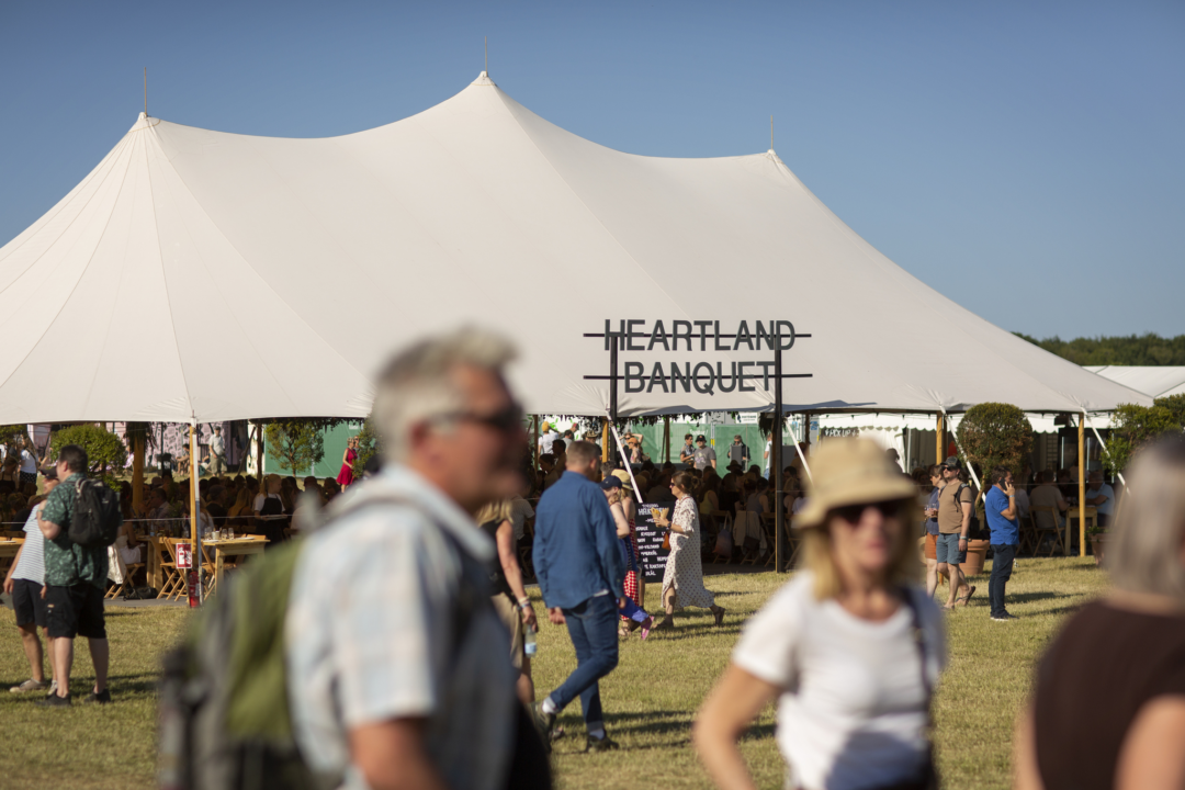 Skiltet 'Heartland Banquet' i sort metal rejser sig over festivalgæster og hvide telte i solskin ved Heartland Festival.