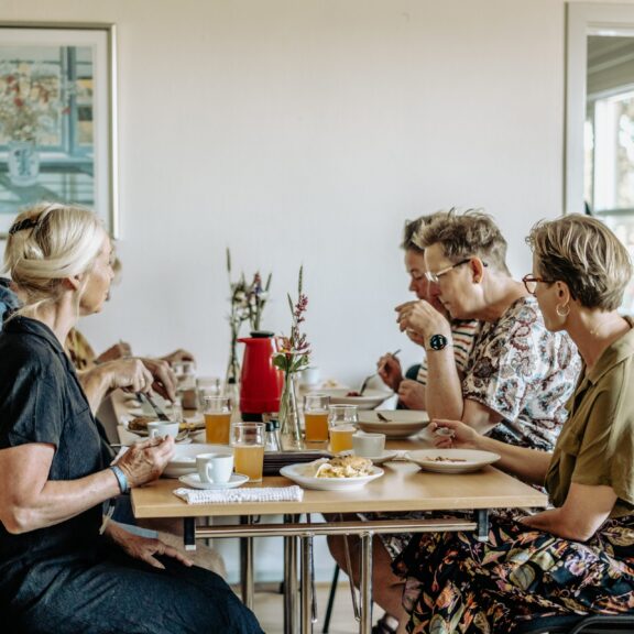 Billedet viser fem voksne personer, der sidder omkring et lille, lyst træbord og spiser et måltid sammen i et roligt, hjemligt miljø.