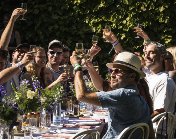 Glade festivalgæster skåler med hvidvin ved et smukt dækket langbord med blomster foran en grøn hæk ved Heartland Festival.