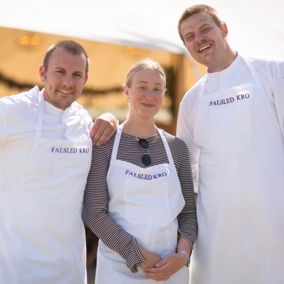 Billedet viser tre voksne personer, der står tæt sammen udendørs i sollys og smiler mod kameraet. De har hvide forklæder på med teksten “Falsled Kro” trykt på brystet.