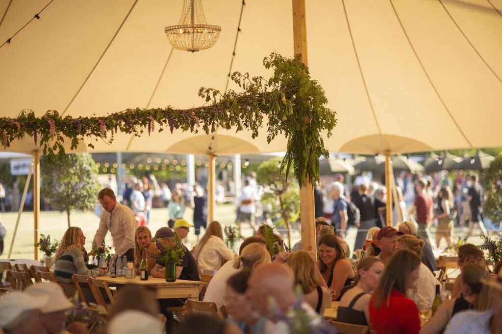 Gæster sidder ved træborde under et smukt dekoreret telt med krystallysekrone og grønne blomsterguirlander ved Heartland Festival.