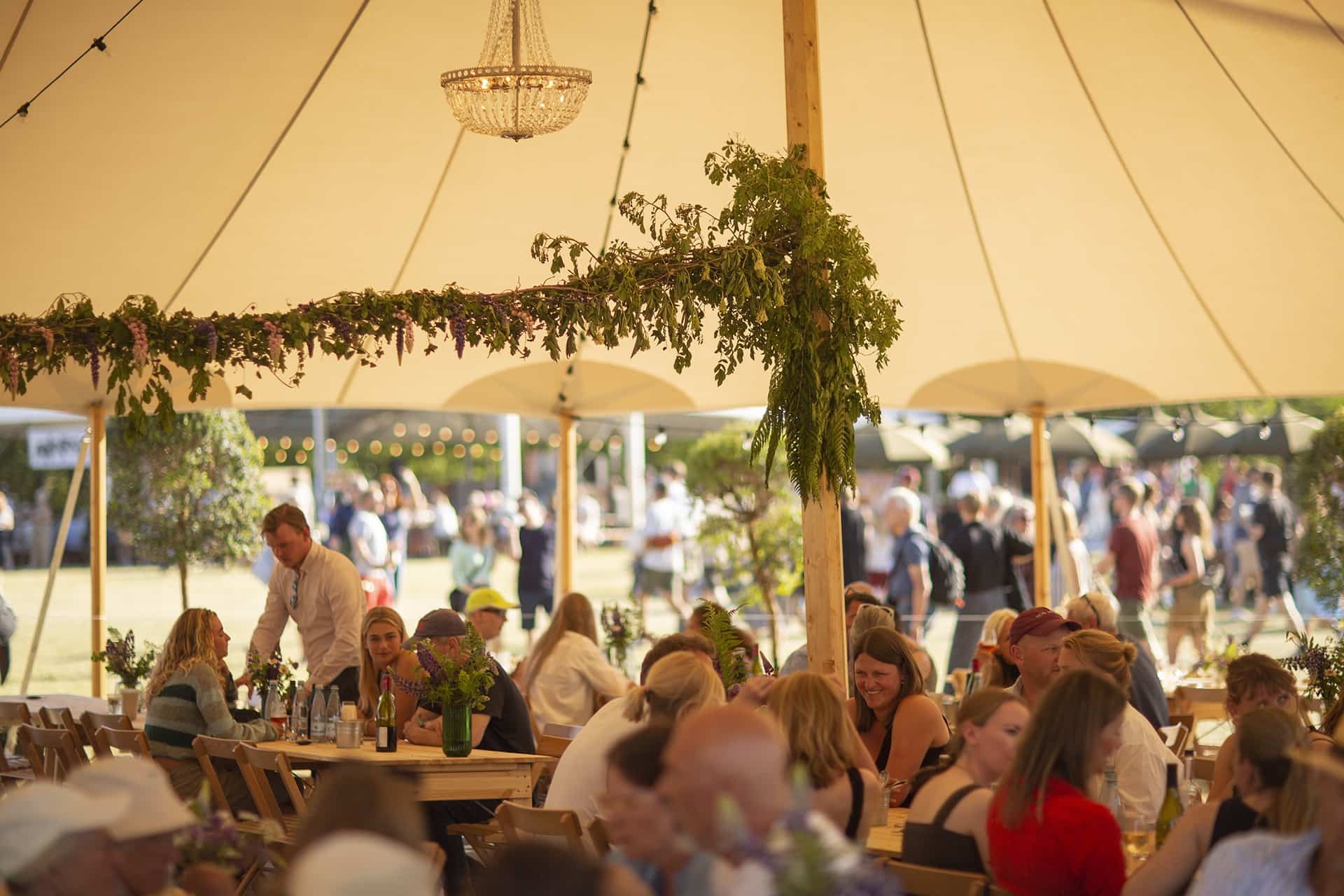 Gæster sidder ved træborde under et smukt dekoreret telt med krystallysekrone og grønne blomsterguirlander ved Heartland Festival.