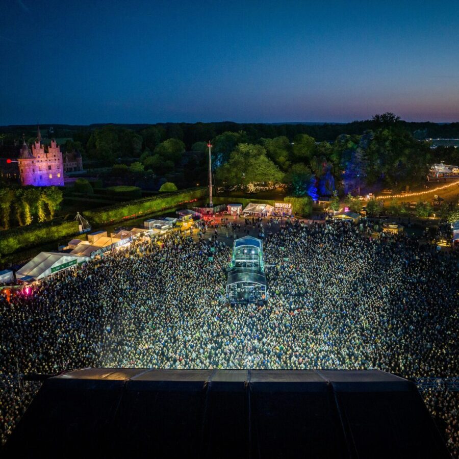 Dronefoto af Heartland Festival i skumringen set fra scenen. Tusindvis af festivalgæster fylder pladsen foran hovedscenen, med Egeskov Slot oplyst i lilla i baggrunden og festivalområdets telte og lys spredt mellem de grønne træer.