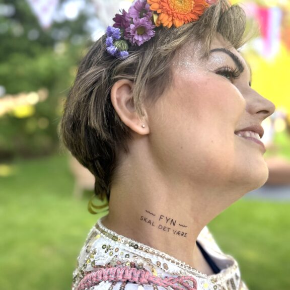 Billedet viser en kvinde som står tæt på kameraet og viser hendes hals frem, hvor der på siden er tatoveret "Fyn skal det være". Kvinden har kort hår med en farverig blomsterkrans og sølv glimmer ved øjenlåget.