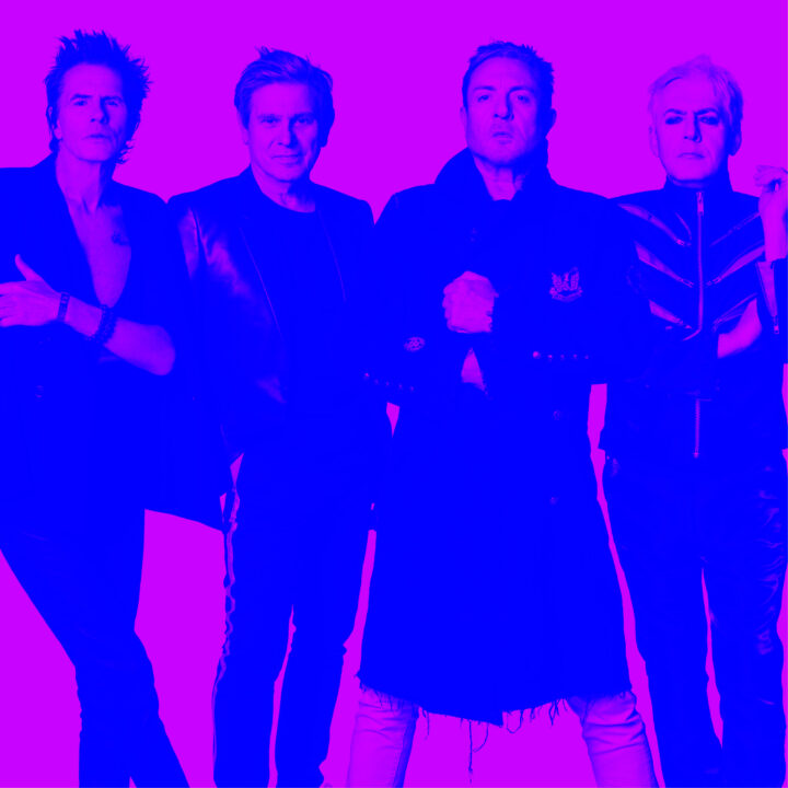 Billedet viser fire voksne mænd (Duran Duran), der står side om side og vender frontalt mod kameraet. De står tæt sammen i en selvsikker positur. Hele billedet er farvet i kraftige neonfarver – en intens pink/magenta i baggrunden og en dyb blå farvetone hen over personerne, hvilket giver et dramatisk og stiliseret udtryk.
