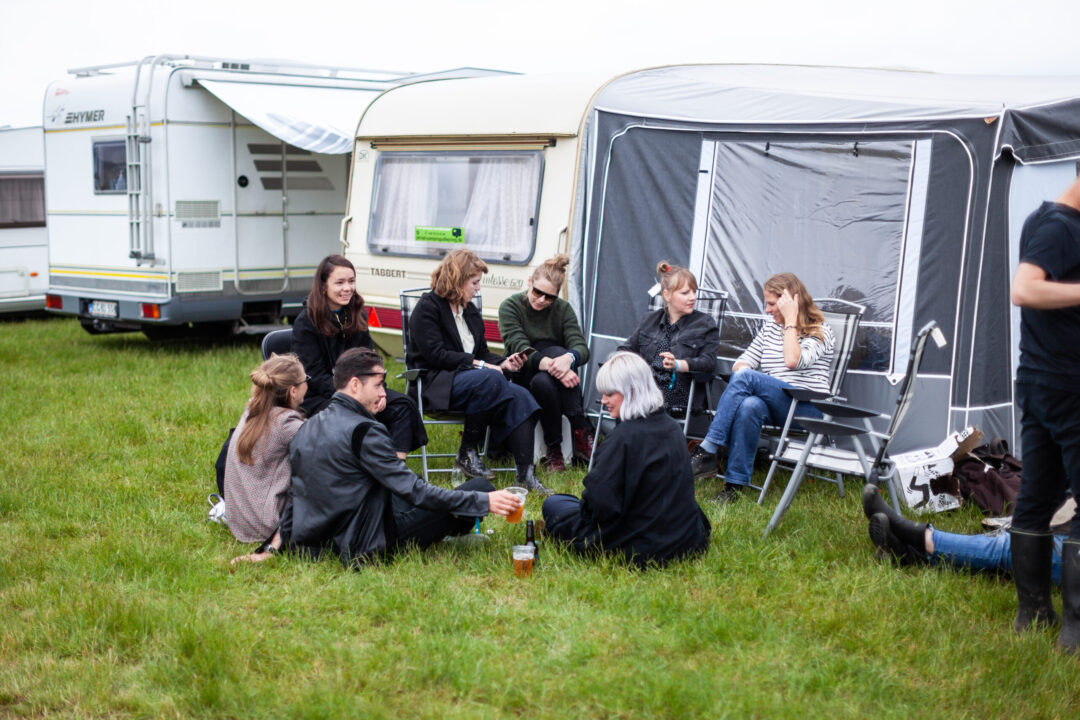 En gruppe mennesker, der sidder i campingstole og på grønt græs foran deres campingtelt. Der er god stemning og rosévin i vinglas.