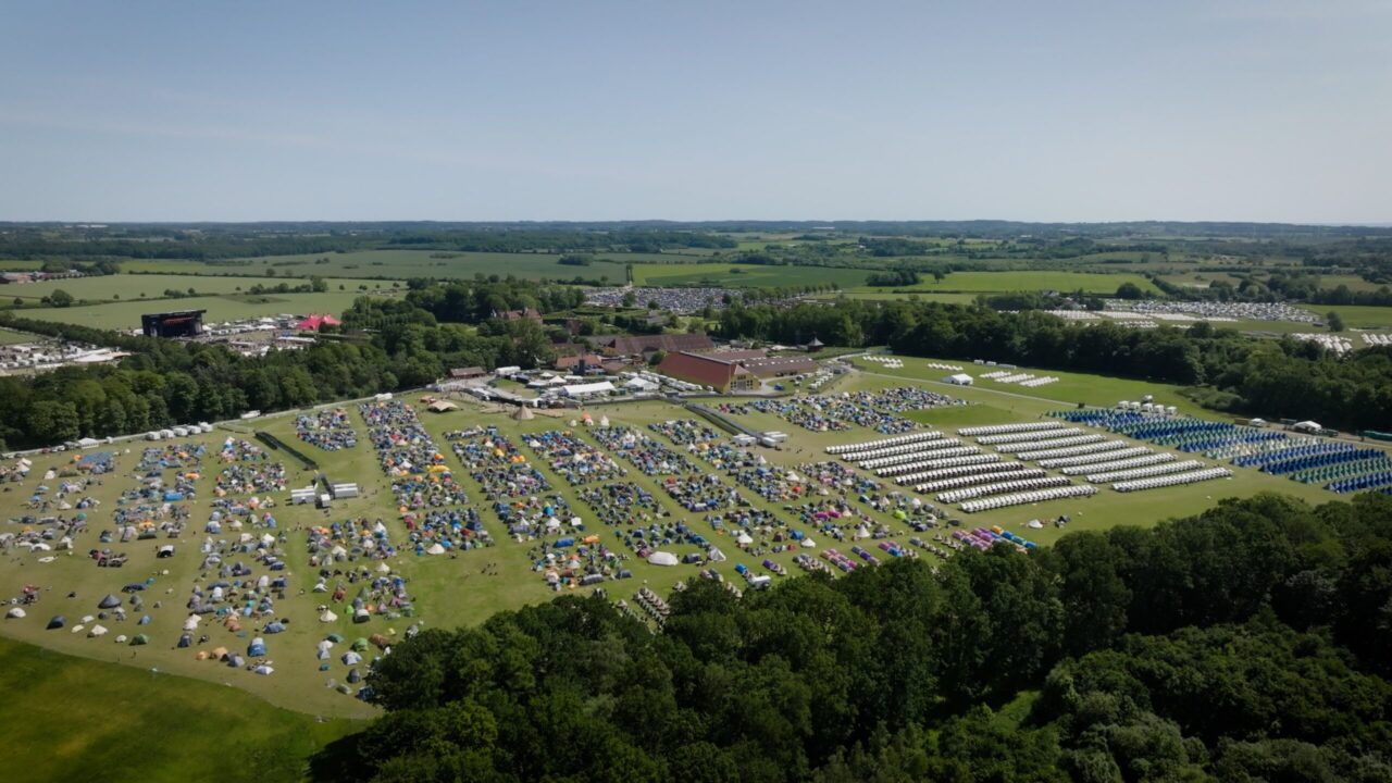 Dronebillede fra campingområdet med en masse opslåede telte. Græsset er grønt og himlen er blå.