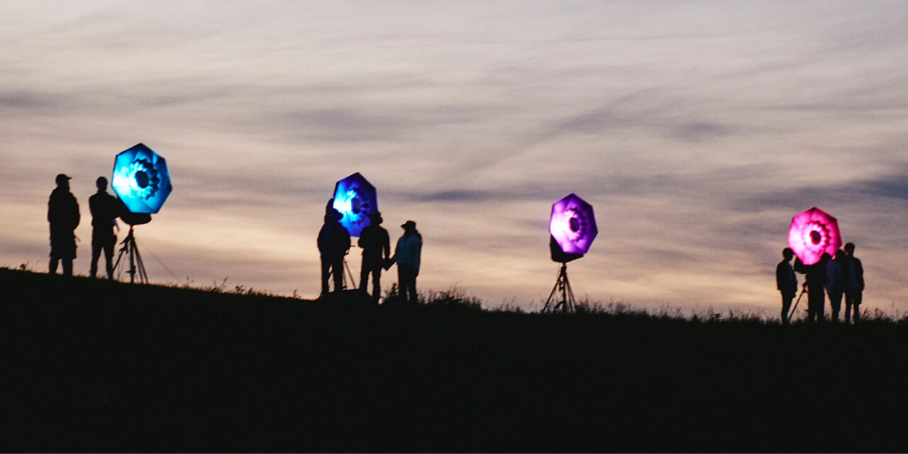 Kunstværk under åben aftenhimmel. 4 lamper med blåt, lilla og pink lys i.