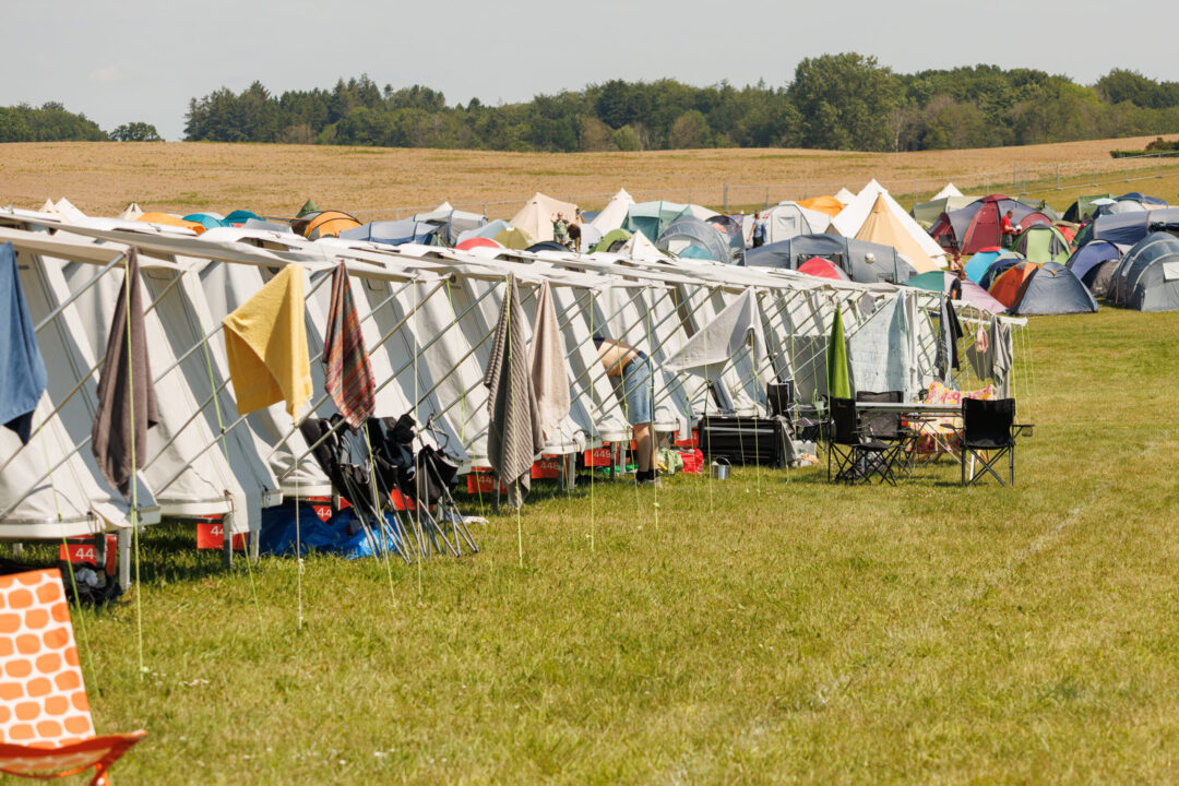 Mane Tent House's på række, hvor der hænger håndklæder på markisen. Man kan se der bor mennesker under festivalen. De står på en grøn græsplæne med marker i baggrunden.