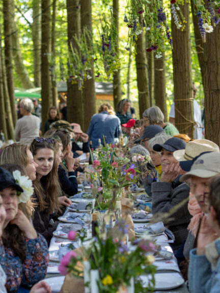 Mennesker som sidder i blandt træerne i skoven ved langbordsmiddag og spiser, drikker og taler sammen. På bordet er flotte farverige blomsterbuketter.