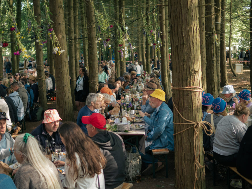 Mennesker som sidder i blandt træerne i skoven ved langbordsmiddag og spiser, drikker og taler sammen.