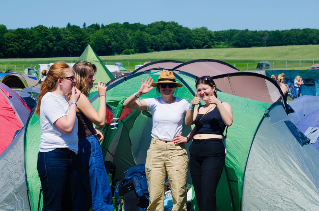 En gruppe piger står og smiler foran deres telt på campingområdet. Himlen er blå, og stemningen er god.