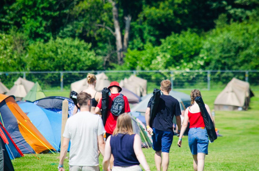 En flok mennesker går med ryggen til på campingområdet og bærer campingstole på ryggen. Græsset og træerne i baggrunden er grønt, og telte står opslået omkring dem.