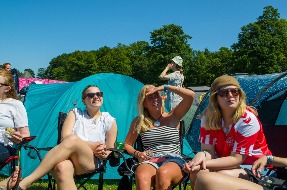 Tre glade piger sidder i campingstole på teltområdet. De har sommertøj på, og solen skinner.