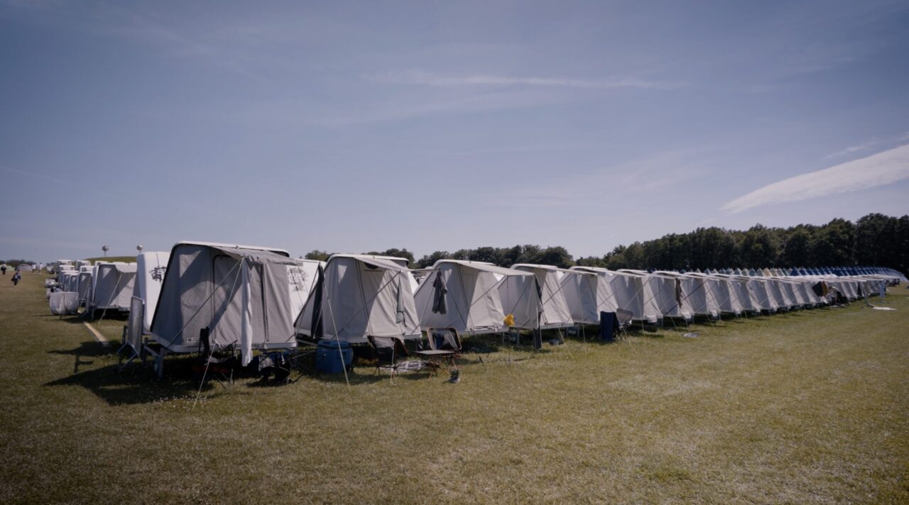 Flere grå tenthouses står på række, og nogle af dem hænger håndklæder til luftning på overdækningen. Himlen er blå over tenthouseområdet.