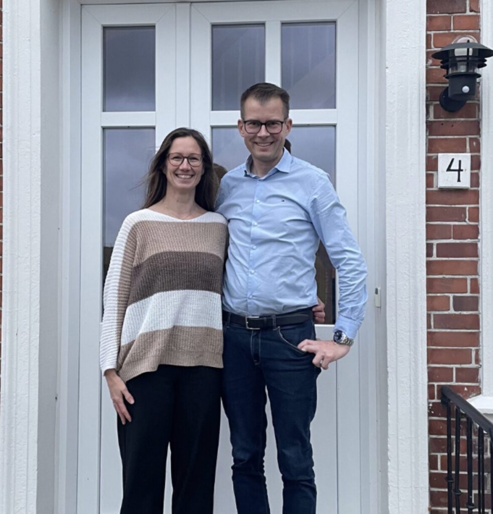 Et billede af et par, der smiler foran en hoveddør i et rødt murstenshus