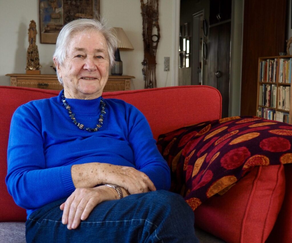 En ældre sød kvinde, der sidder i sin røde sofa i hendes stue. Hun ser mild og venlig ud.