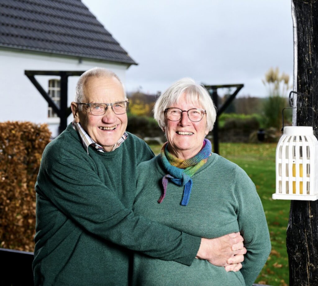 Et sødt ægtepar i grønne striktrøjer, der står foran deres hjem. De smiler og han holder om hende.