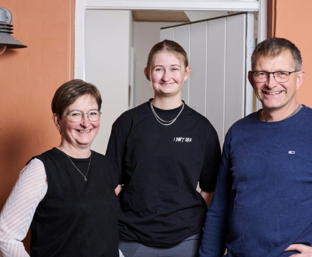 En far, mor og datter i midten. De står smilende i deres hoveddør og ser velkomne ud. Deres hus er orange.