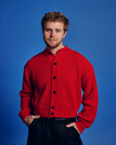 Alex Warren iført rød sweater står foran blå baggrund.